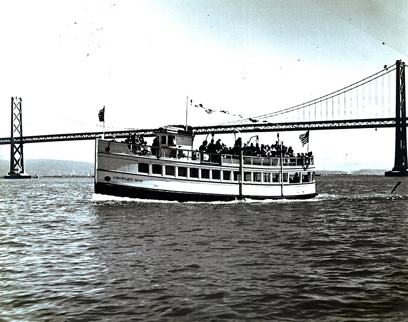 A vintage black and white photo of a 1939 Crowly Marine Red and White Fleet sightseeing boat crowded with passengers on the San Francisco Bay, with a bridge tower in the background.