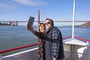 A happy couple taking a selfie on a Red and White Fleet bay cruise with the Golden Gate Bridge behind them.
