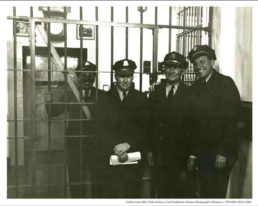 four Alcatraz correctional officers in formal uniforms standing guard at the iron-barred Sallyport entrance during the federal penitentiary era.