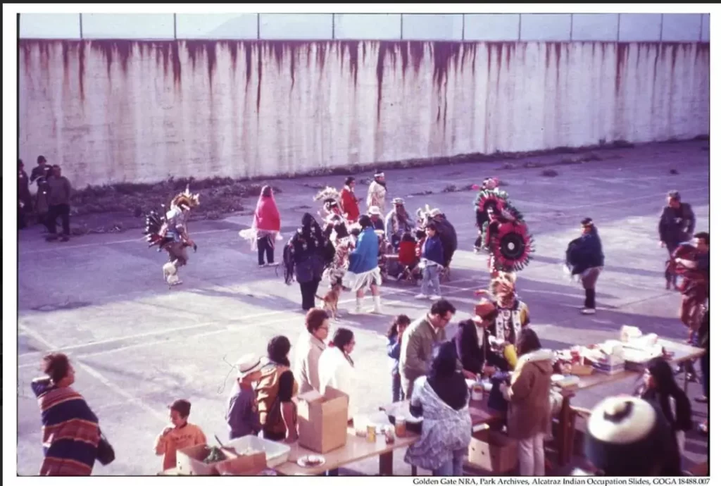 historical photo of Native American activists, including men, women, and children, gathered around a large table sharing a traditional Thanksgiving feast during the 1969 occupation of Alcatraz Island.