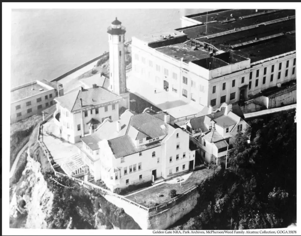 A historical aerial photograph showing the 15-room Warden's House mansion and the adjacent 84-foot reinforced concrete Alcatraz Lighthouse on the island’s summit during the 1940s.
