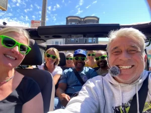 Tour guide Jo-Jo and six guests taking a smiling group selfie inside an open-air Jeep during a San Francisco sightseeing tour