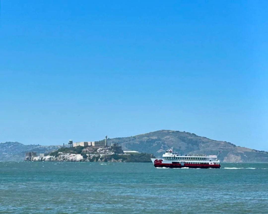 A Red and White Fleet sightseeing boat cruising past the historic buildings and lighthouse of Alcatraz Island in San Francisco Bay.