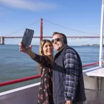 A happy couple taking a selfie on the deck of a Red and White Fleet boat with the Golden Gate Bridge and San Francisco Bay in the background.
