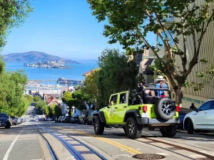 Tour guests in an open-air Jeep looking down a San Francisco cable car line with Alcatraz visible in the distance